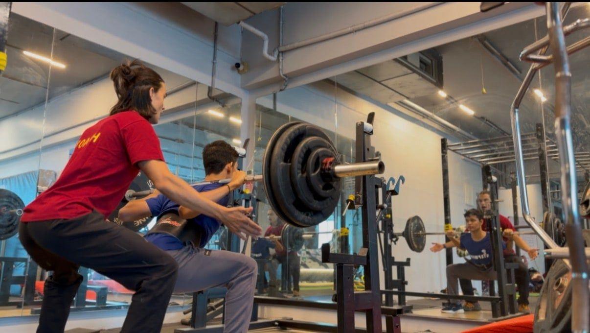 A coach supporting a young athlete training at the squat rack